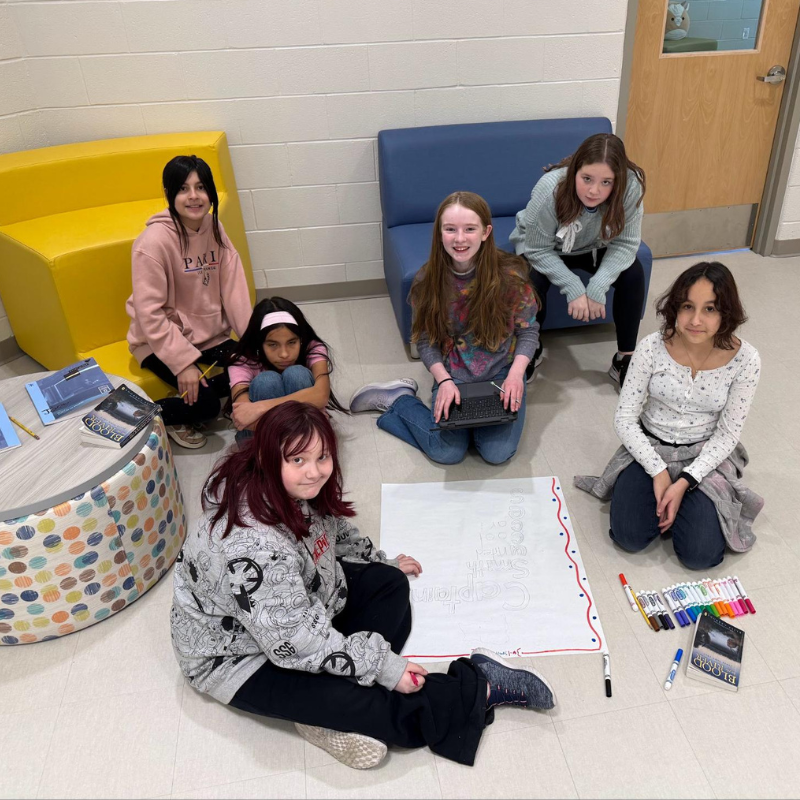 Five young people collaborate on a project, sitting on the floor around a large poster.