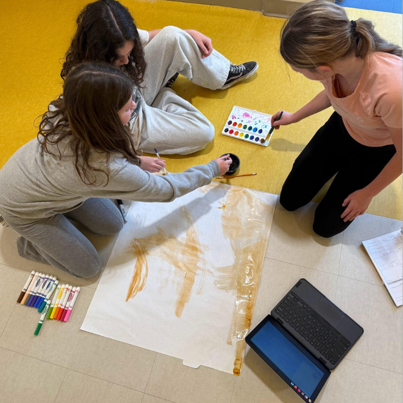 Three young people are painting on a large sheet of paper on the floor.