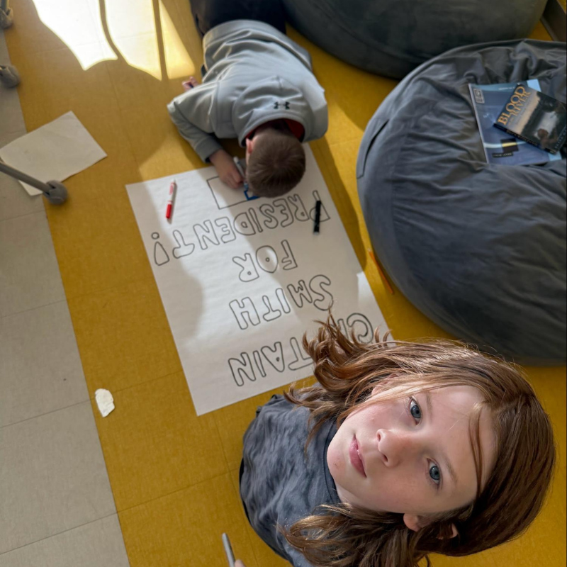 Two children work on a poster that reads 'CAPTAIN SMITH FOR PRESIDENT!' on a yellow floor.