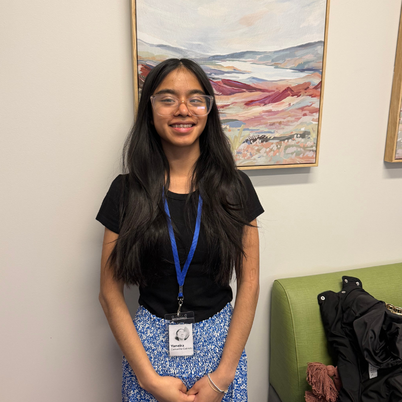 A young woman with long dark hair and glasses smiles, wearing a black shirt and a blue patterned skirt.