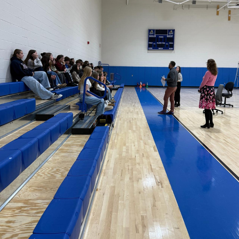 Students sit on blue bleachers in a gymnasium, facing a group of adults.