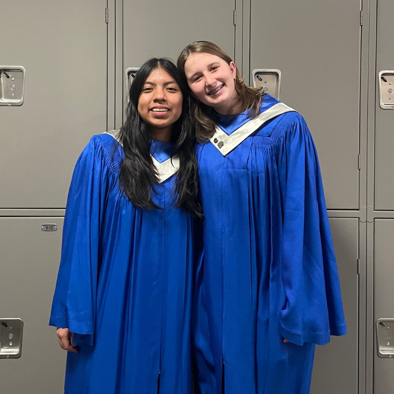 Two smiling young women in matching blue choir robes stand in front of gray lockers.