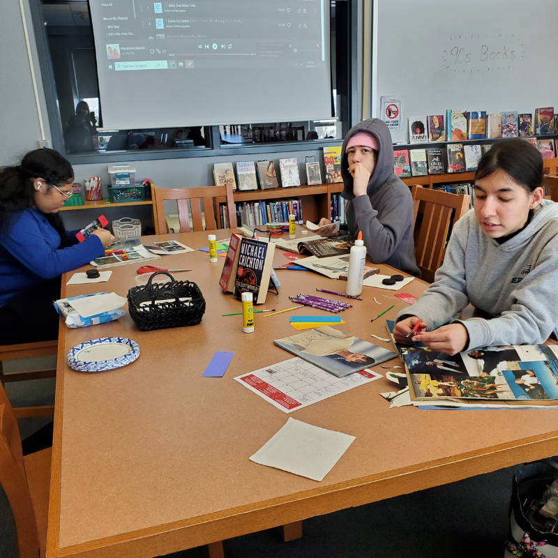 Three young people are gathered around a table, cutting and pasting images from magazines.