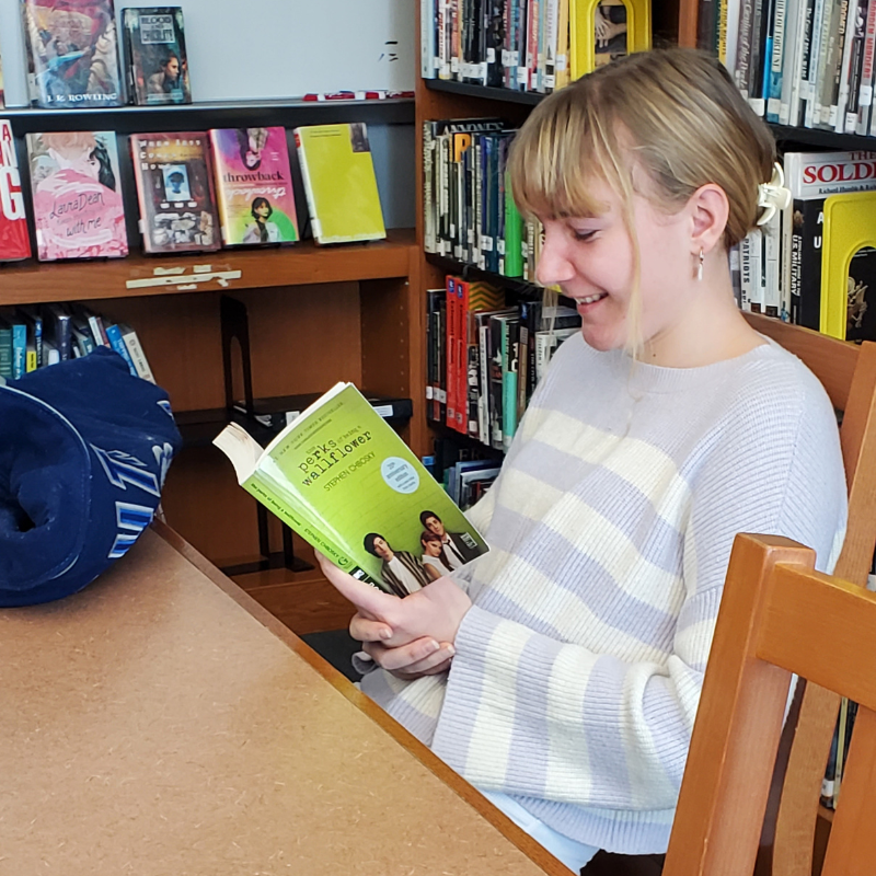 A young woman smiles while reading 'The Perks of Being a Wallflower' in a library.