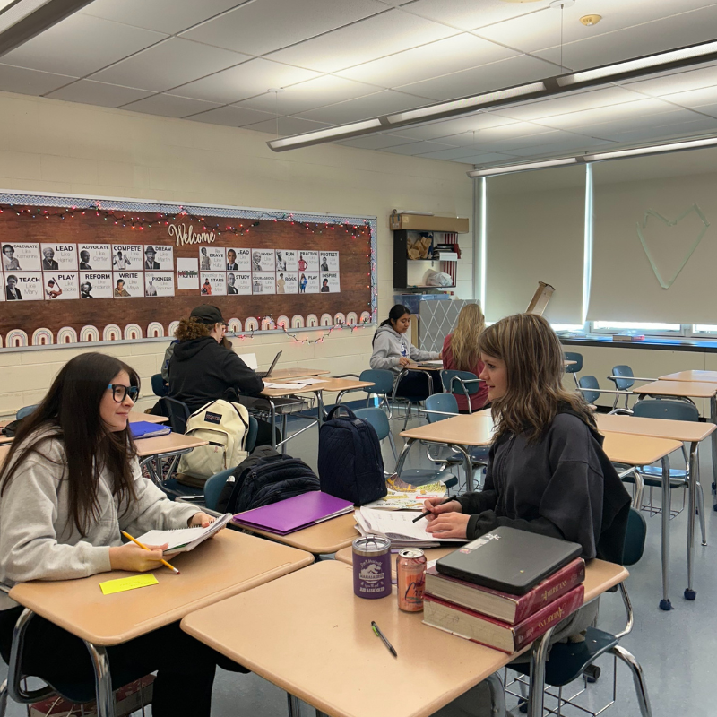Students work at desks in a classroom, with a bulletin board displaying inspirational words and figures.