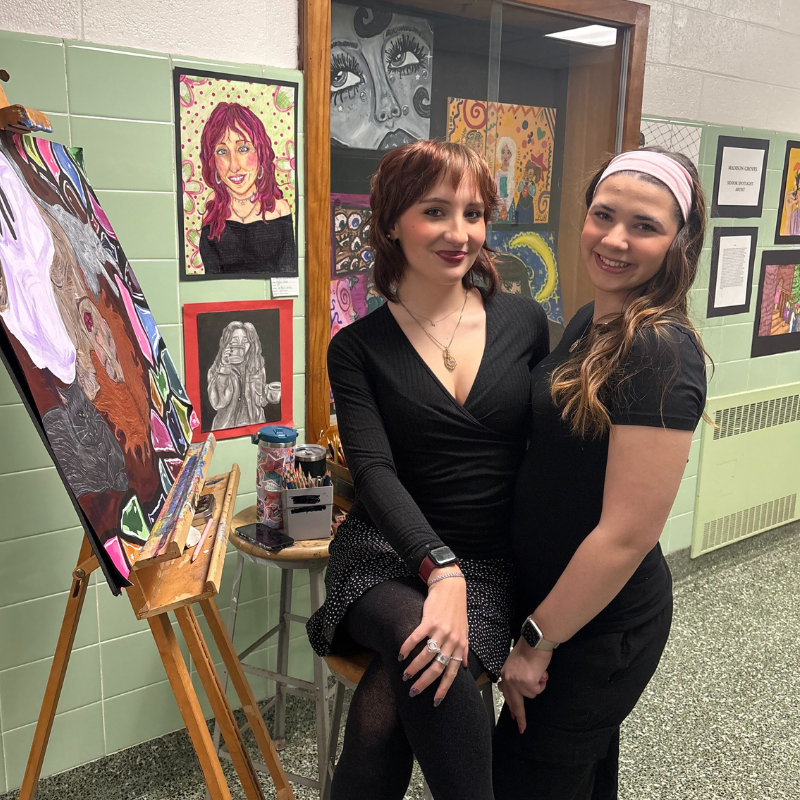 Two young women smile in front of a display of colorful artwork, including a large painting on an easel.