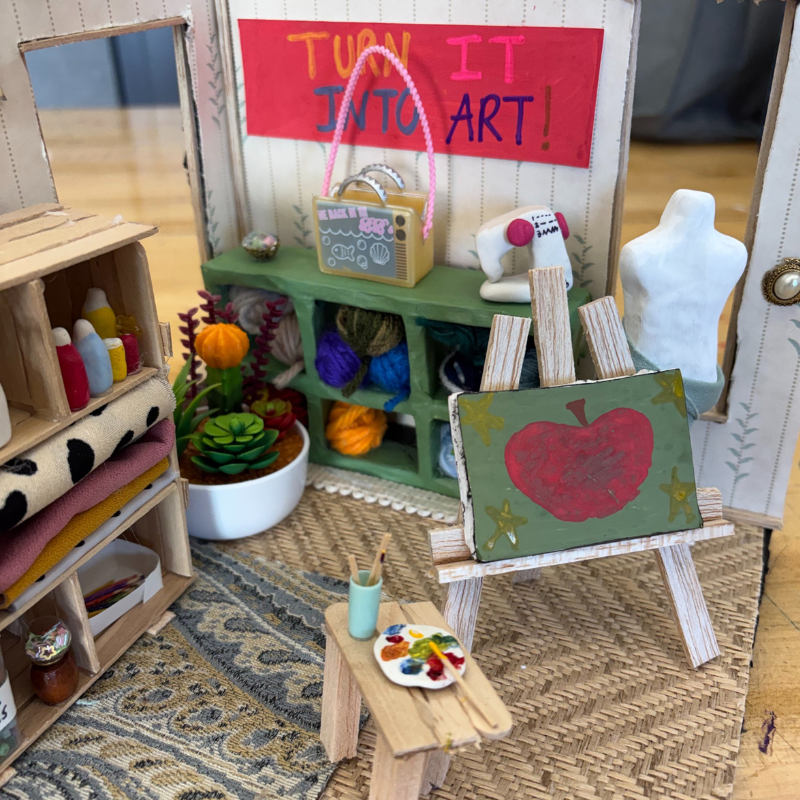 A miniature art studio with a painting of an apple on an easel, yarn, and craft supplies.