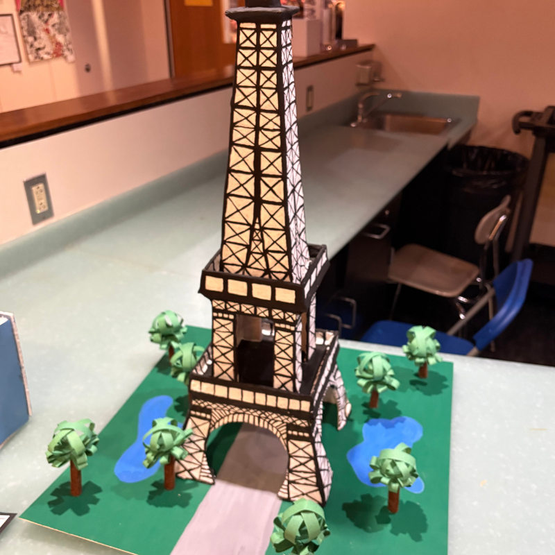 A detailed model of the Eiffel Tower stands on a green base with miniature trees and blue ponds.