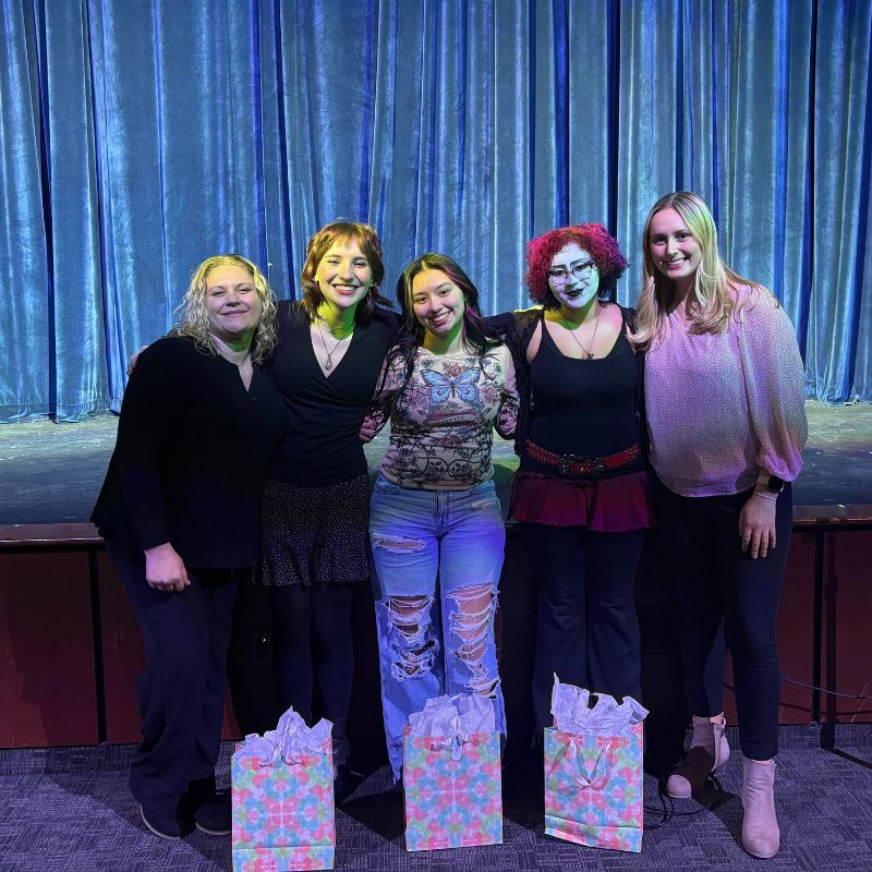Five women stand together in front of a blue stage curtain, smiling.