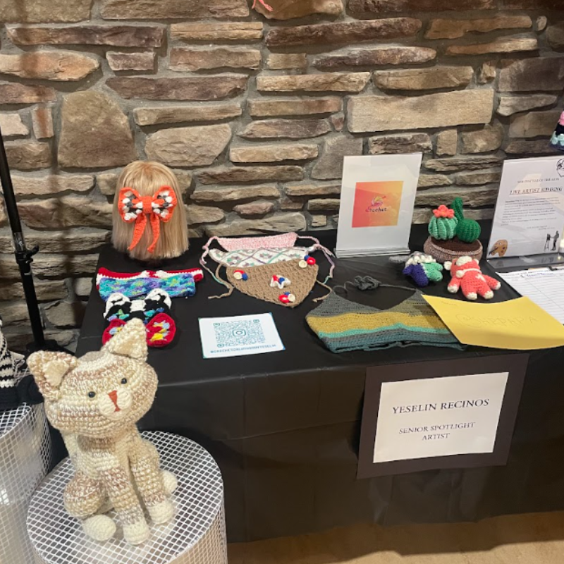 A display table showcases various crocheted items, including a cat, cacti, and face masks, against a stone wall.