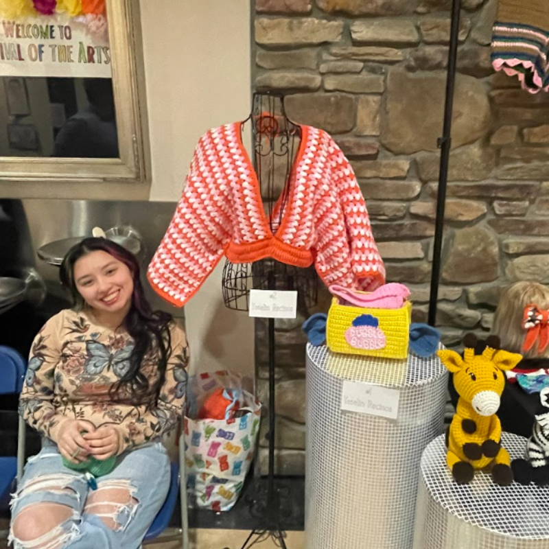 A young woman smiles while sitting next to a display of crocheted items at an art festival.
