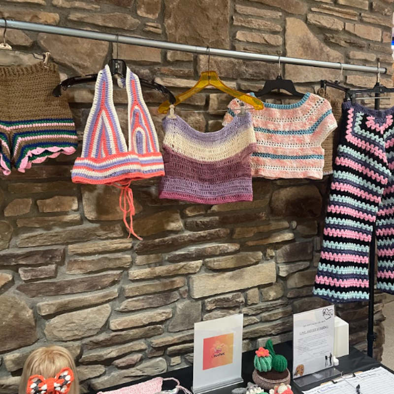 A display of colorful, handmade crochet tops and pants hangs on a rack against a stone wall.