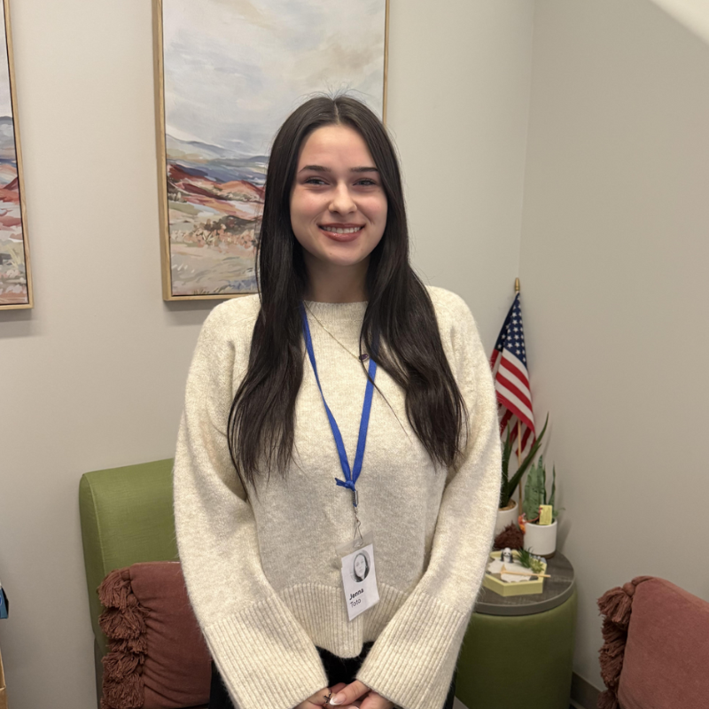 A young woman with long dark hair smiles, wearing a cream-colored sweater and a blue lanyard with an ID badge.