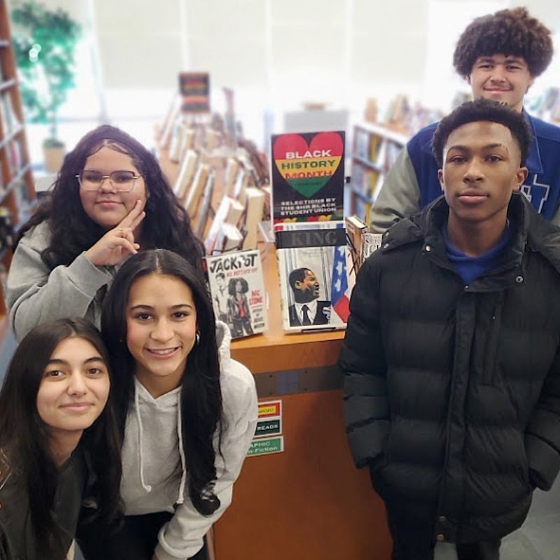 Five students stand in front of a bookshelf displaying Black History Month selections.
