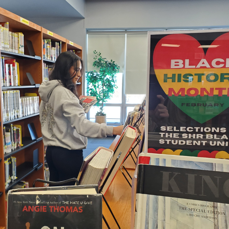 A young woman browses books in a library, with a Black History Month display in the foreground.
