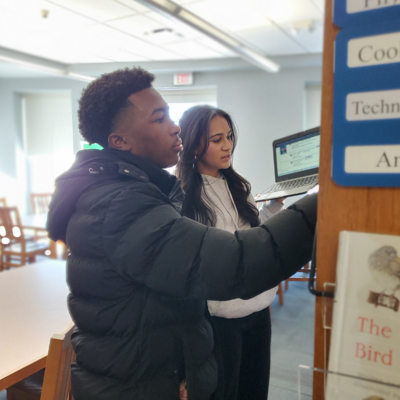 Two students, a boy and a girl, look at a laptop screen in a library setting.