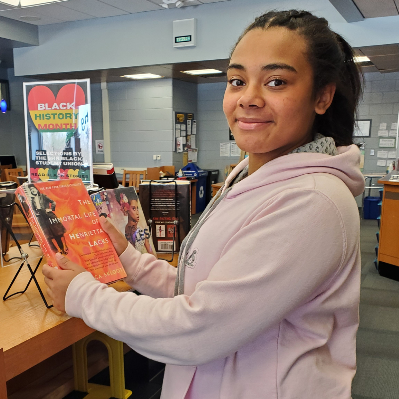 A young person smiles while holding the book "The Immortal Life of Henrietta Lacks" in a library.
