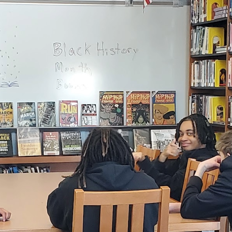 Students in a library setting, with a whiteboard displaying "Black History Month Books" and shelves of books.