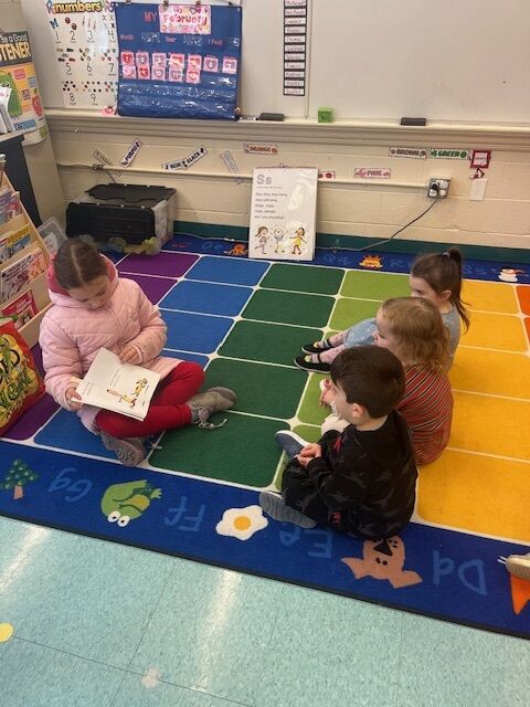 A young girl reads a book aloud to a small group of children sitting on a colorful classroom rug.