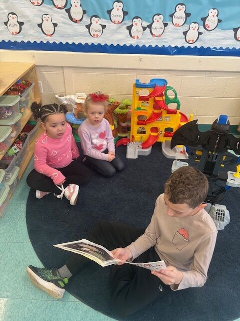 A young boy reads a book to two young girls sitting on a dark blue rug in a classroom.