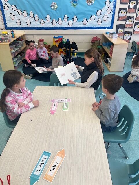 A young girl reads a book to a group of children sitting on the floor in a classroom.
