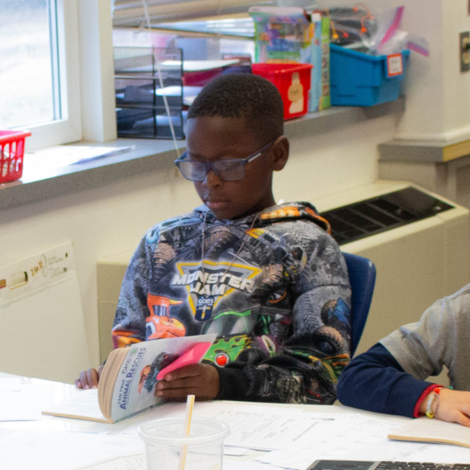 A young boy with glasses reads a book titled "Animal Rescues" in a classroom.