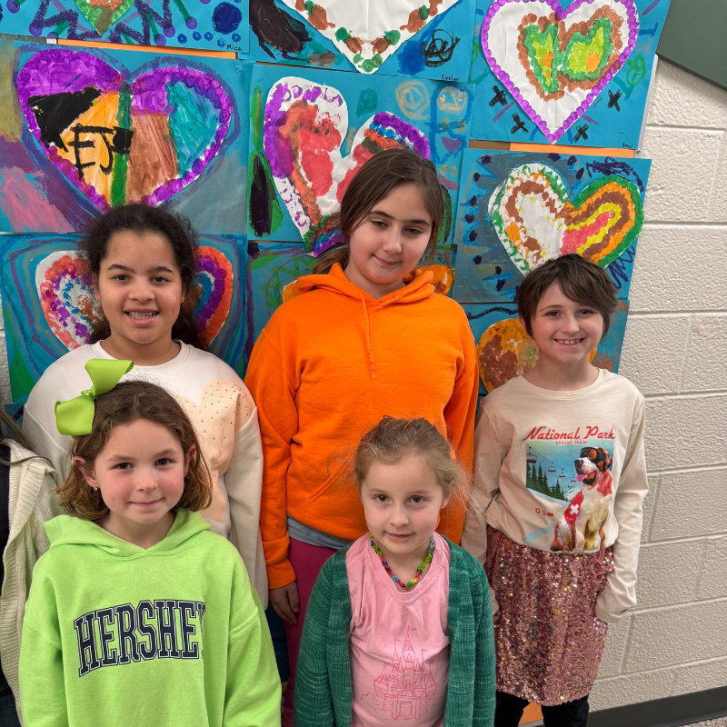 Five smiling children stand in front of a wall decorated with colorful heart-shaped artwork.