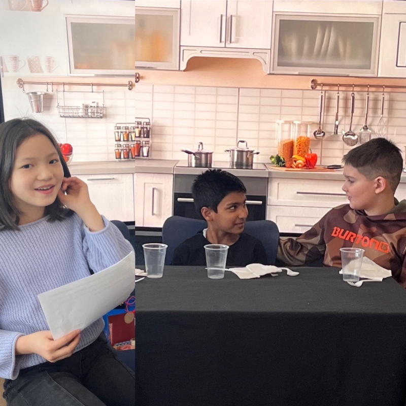 Three children sit at a table in front of a kitchen backdrop, with one girl holding a paper.