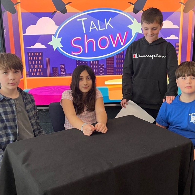 Four children are seated at a table in front of a "Talk Show" backdrop.