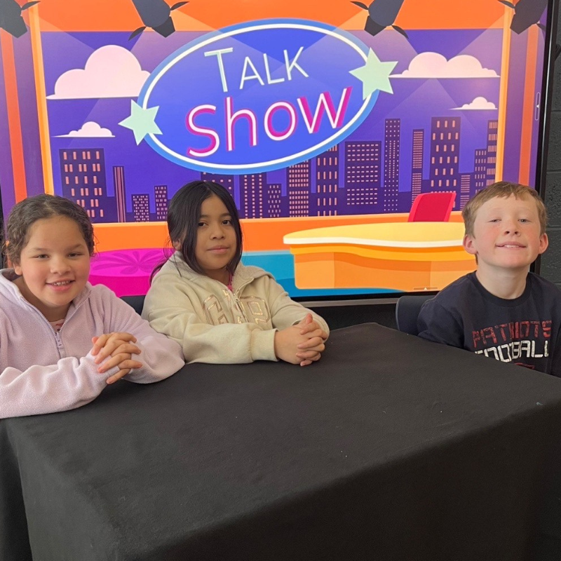 Three children sit at a table in front of a large screen displaying "TALK SHOW" with a city skyline graphic.