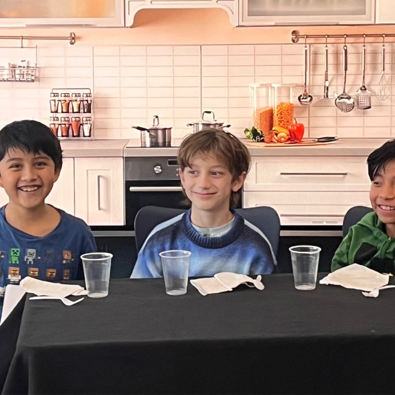Three smiling boys sit at a table in a kitchen, with plastic cups and napkins in front of them.
