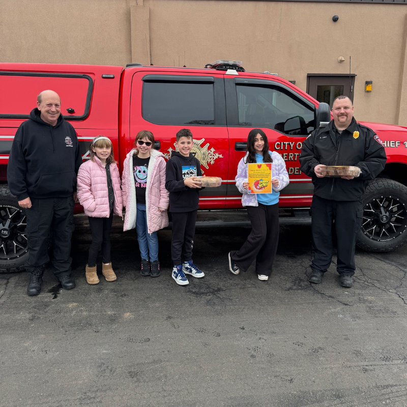 Two firefighters stand with four children in front of a red fire truck, holding baked goods.