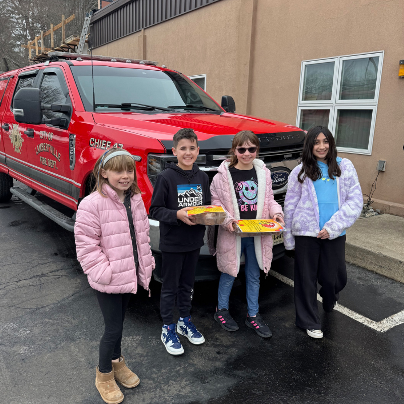 Four children stand in front of a red fire truck, holding boxes of baked goods.