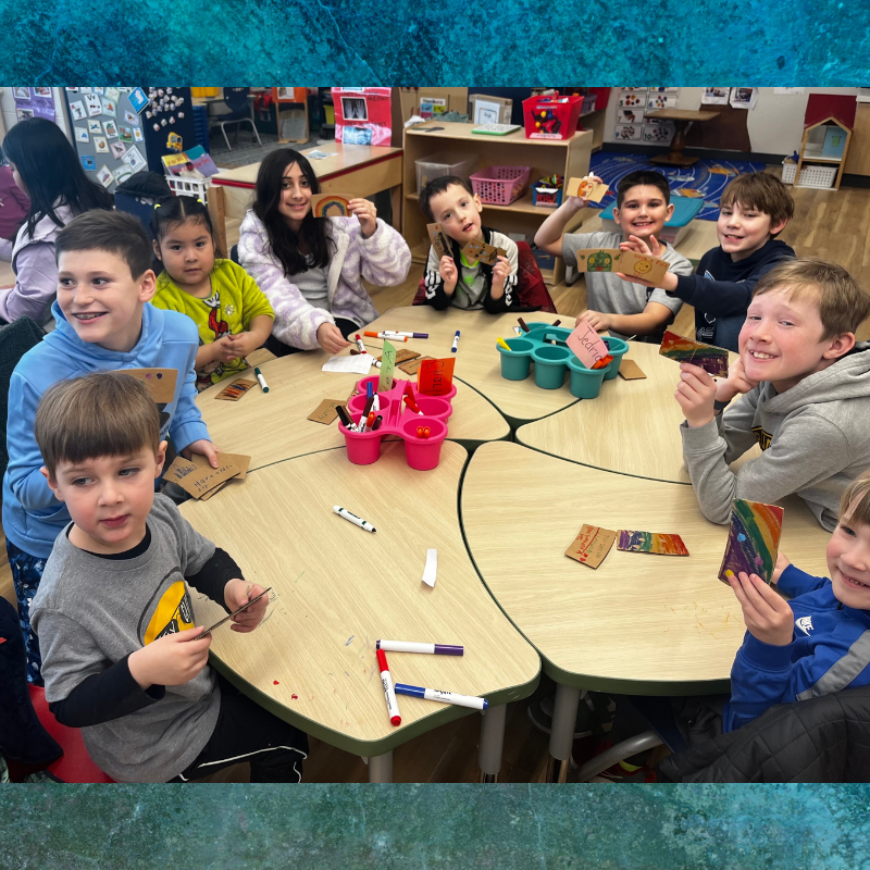 Children at a round table are engaged in an art project, holding up their colorful creations.