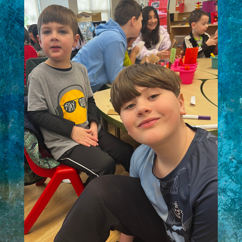 Two young boys smile at the camera in a classroom setting with other children in the background.