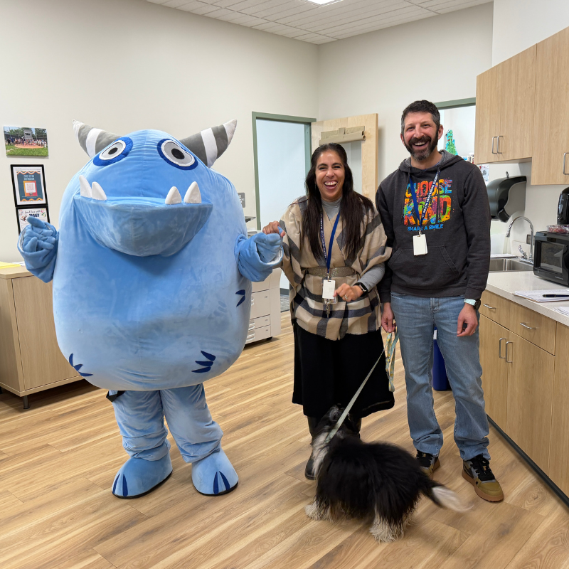A blue monster mascot poses with a woman and a man, while a small dog stands between them.