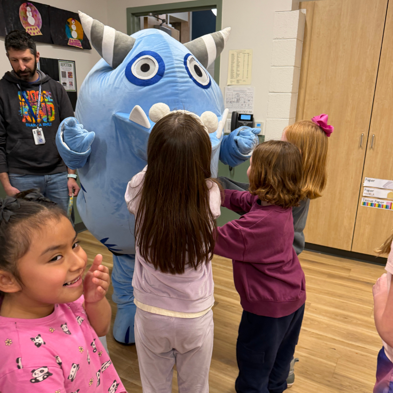 A large, friendly blue monster interacts with a group of young children in a classroom.