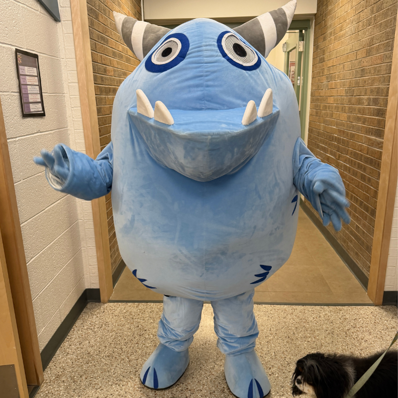 A large, friendly blue monster mascot stands in a hallway, gesturing with its hand.