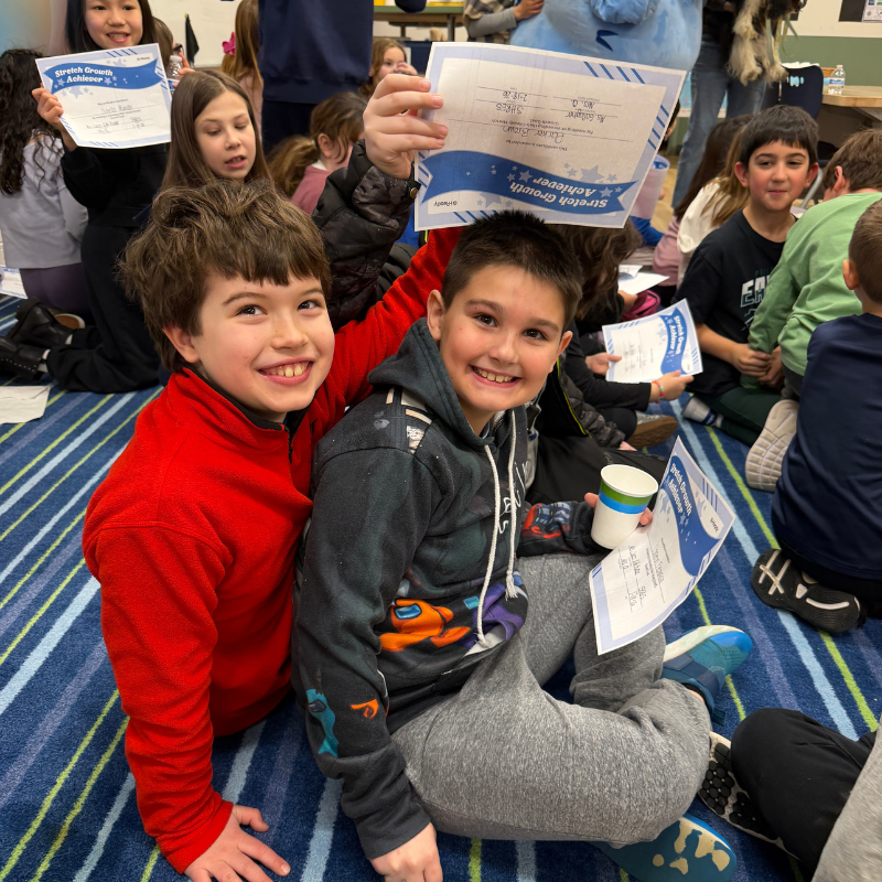 Two smiling boys sit on a striped carpet, holding certificates that say "Stretch Growth Achiever".