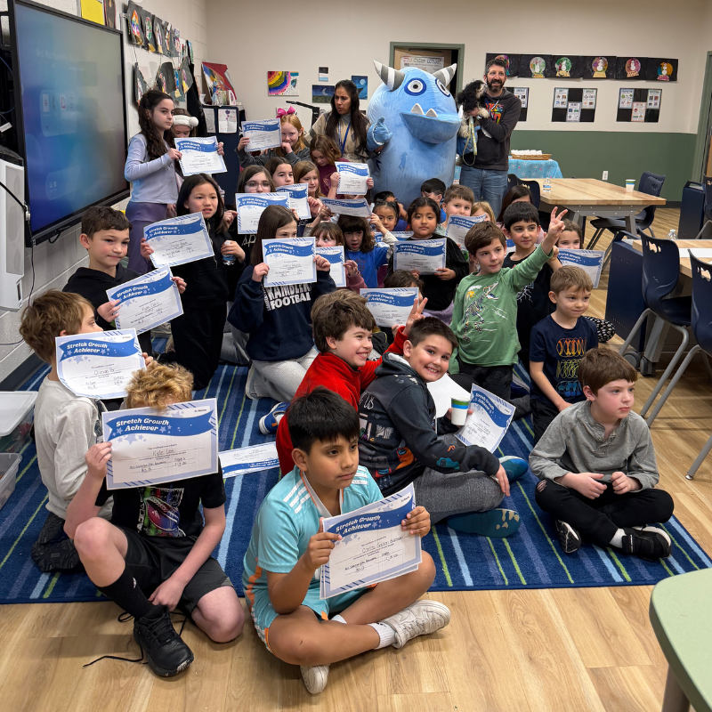 A group of children and adults pose for a photo with a blue monster mascot, holding certificates.