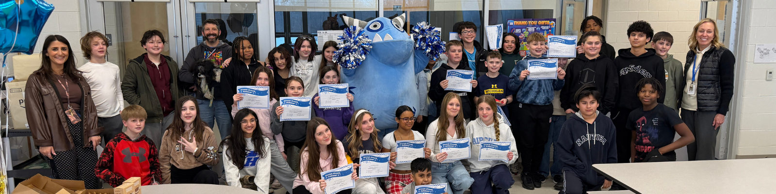 A group of students and adults pose for a photo with a large blue mascot and blue balloons.