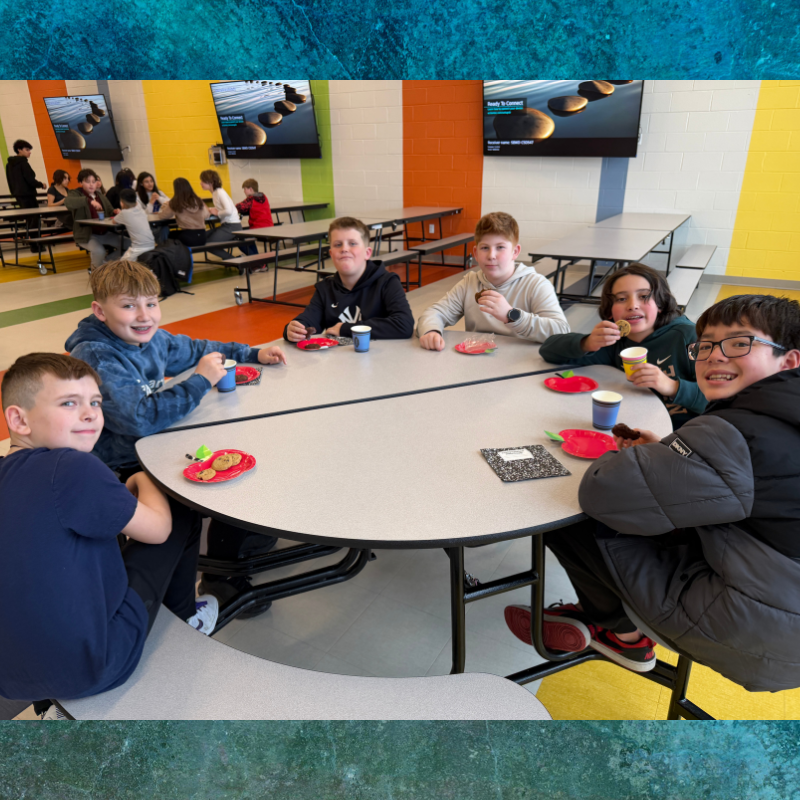 A group of smiling boys sit at a round table in a brightly colored cafeteria, enjoying snacks.