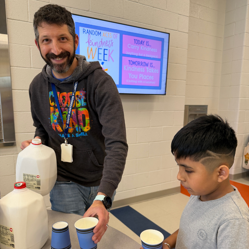 A man pours milk from a jug into cups for a young boy, with a screen displaying "Random Acts of Kindness Week" in the background.