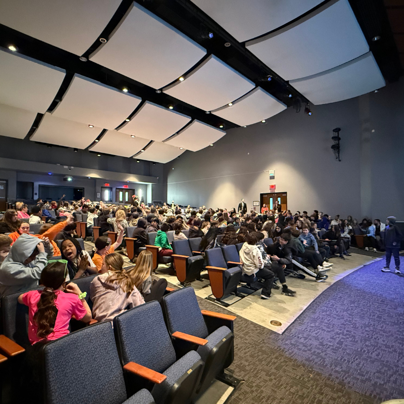 A large audience of students fills an auditorium, seated in rows of blue chairs.