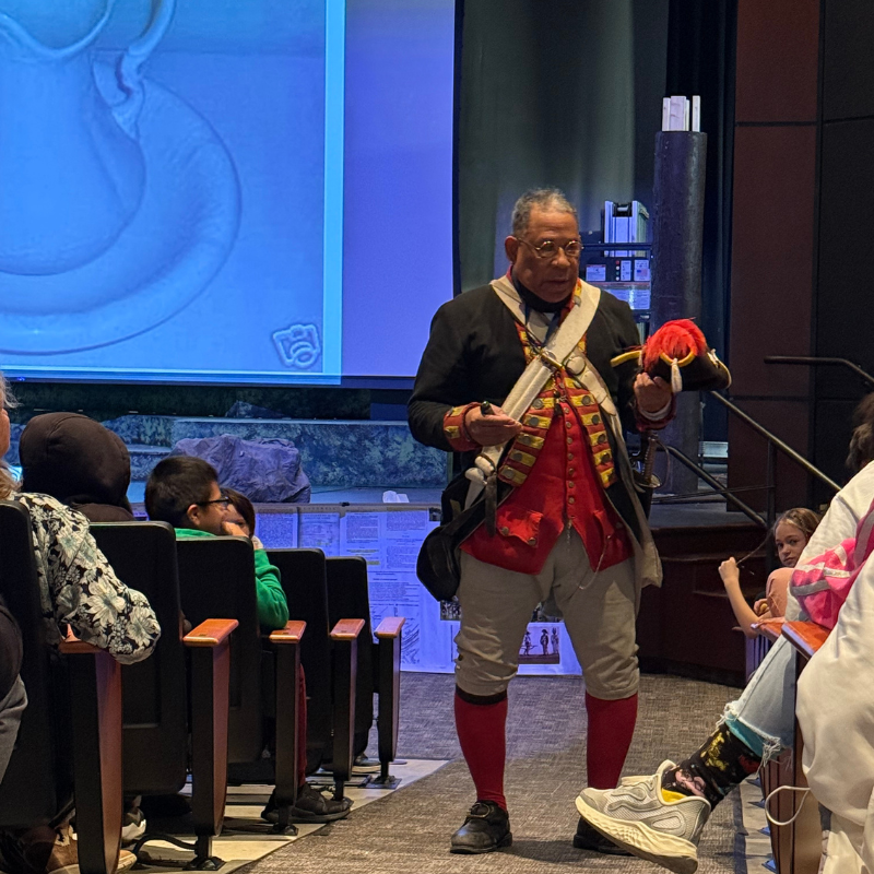 A man in a historical costume, holding a feathered hat, speaks to an audience seated in an auditorium.