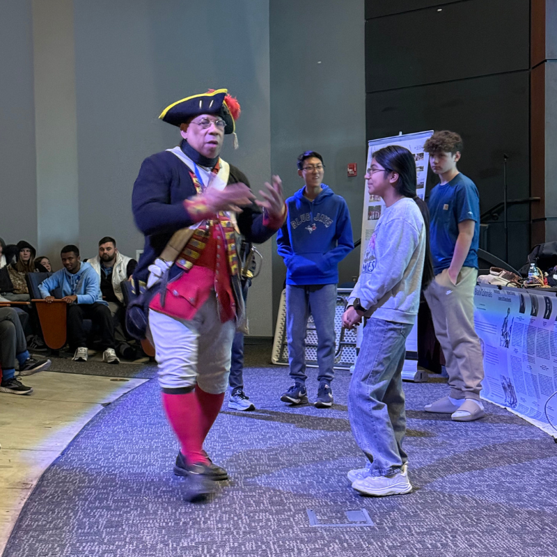 A historical reenactor in colonial attire speaks to a group of students.