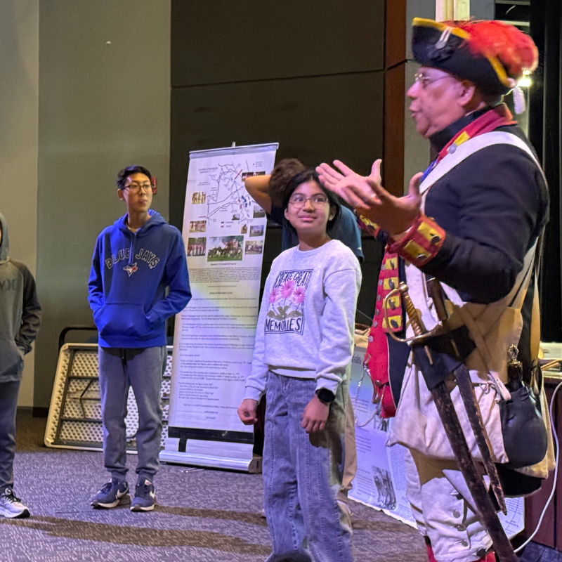 A historical reenactor in a colonial uniform speaks to a group of young people.