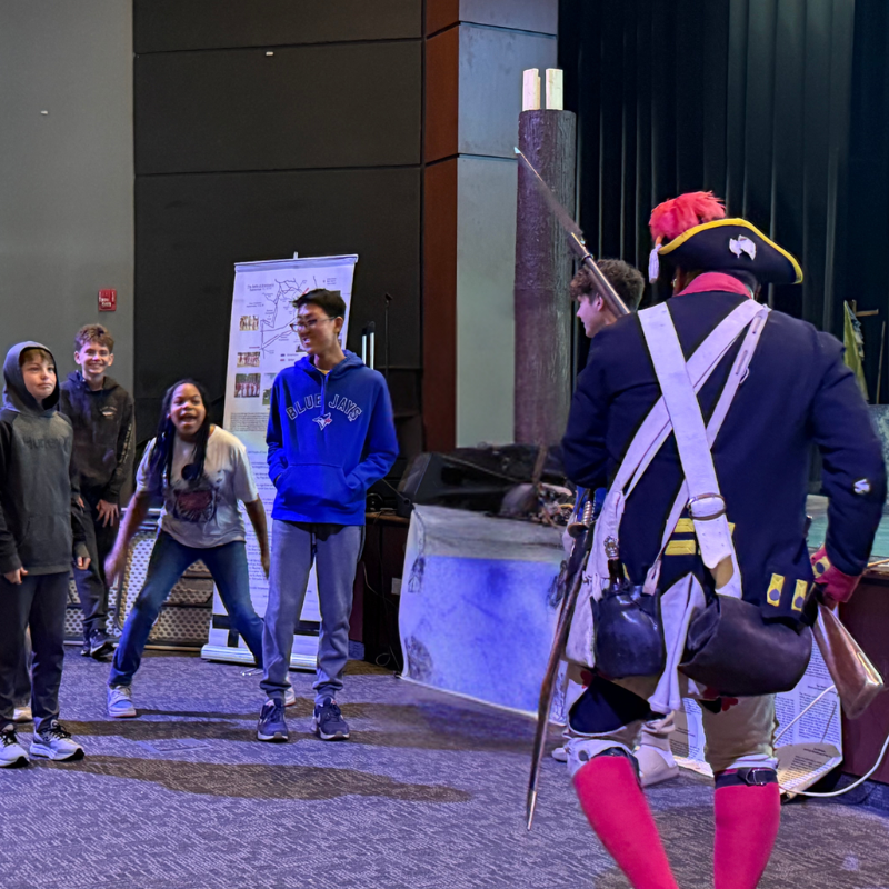 Students watch a historical reenactor dressed in colonial attire holding a musket.