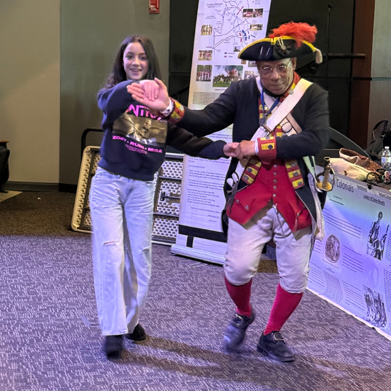 A young woman in jeans and a sweatshirt dances with a man in a historical colonial costume.