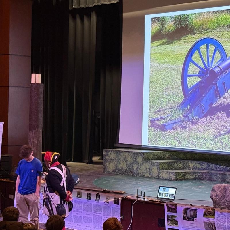 A presenter in historical costume stands on a stage next to a large screen displaying a blue cannon.